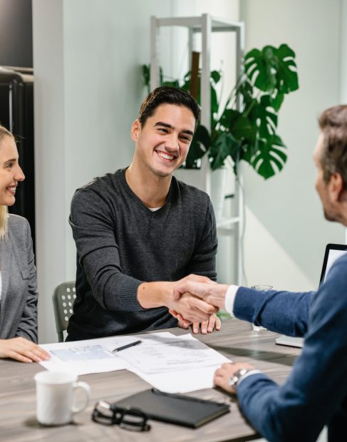 Young happy couple making an agreement with their financial advisor at home. Men are shaking hands.